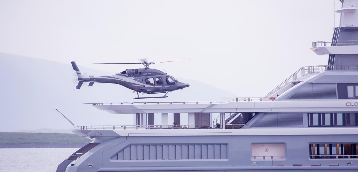Helicopter hovering above the helipad of a large luxury yacht on calm water.
