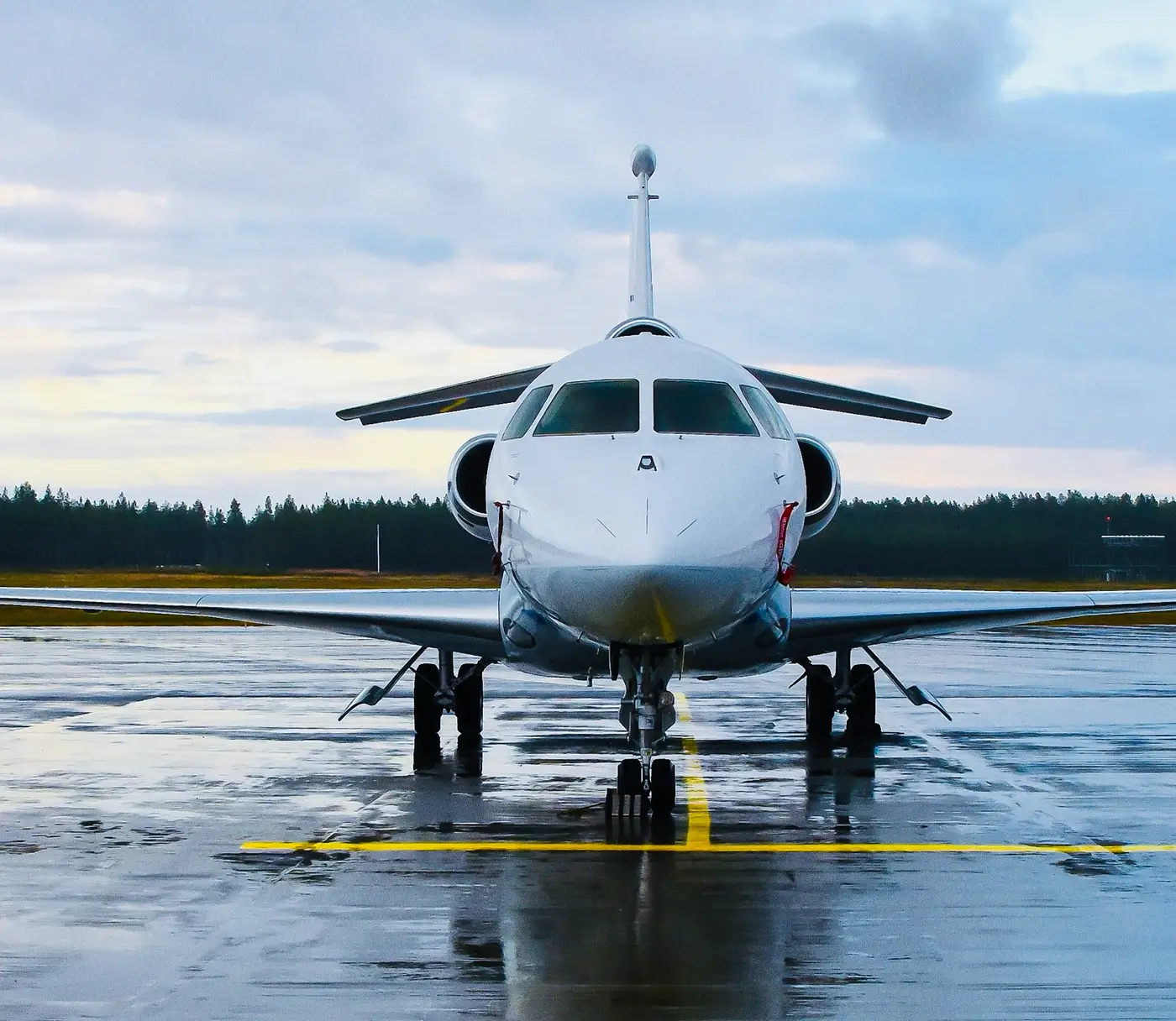 Widok od przodu białego prywatnego odrzutowca zaparkowanego na mokrym pasie startowym, z lasem w tle pod pochmurnym niebem.