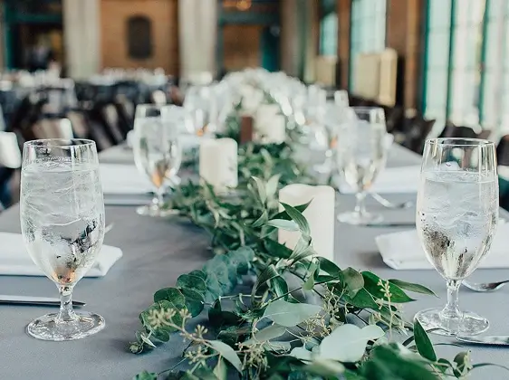 Mesa de comedor larga con vasos de agua, velas blancas y un centro de mesa con hojas verdes en un elegante espacio para eventos.