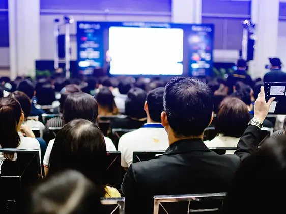Audiencia de personas sentadas frente a un escenario con una pantalla grande y brillante en una conferencia o seminario.