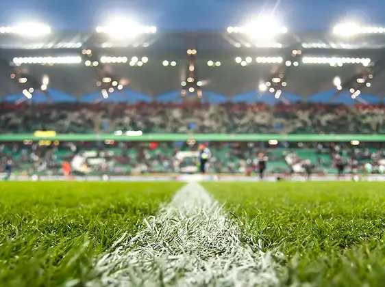 Línea blanca sobre césped verde en un estadio con multitud borrosa y luces brillantes de fondo.