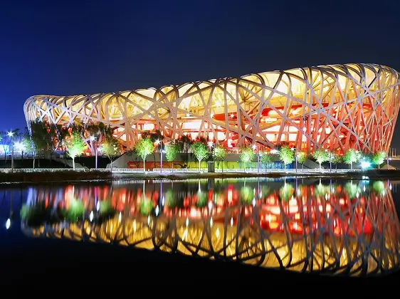 Vista nocturna del Estadio Nacional de Beijing iluminado en amarillo y rojo, reflejado en el agua cercana.