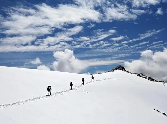 Cuatro excursionistas suben por una cresta montañosa cubierta de nieve bajo un cielo azul con nubes dispersas.