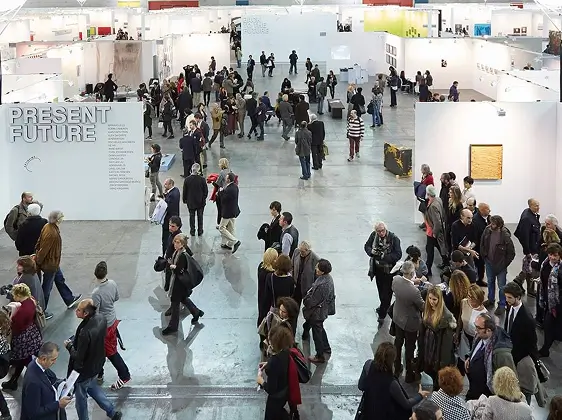 Multitud de personas caminando e interactuando dentro de una gran sala de exposiciones de arte moderno con paredes blancas y un letrero que dice «FUTURO PRESENTE».