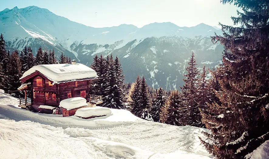 Capanna di legno coperta di neve accanto ai pini in un paesaggio invernale montuoso sotto un cielo limpido.