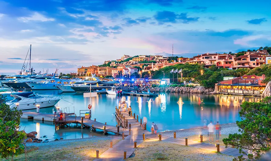Scenic marina at dusk with yachts docked, boardwalk, and illuminated buildings on a hill.