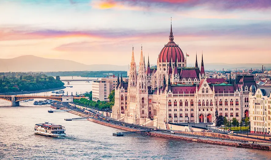 Vue panoramique sur le bâtiment du Parlement hongrois au bord du Danube au coucher du soleil avec un bateau de croisière fluvial passant à Budapest.