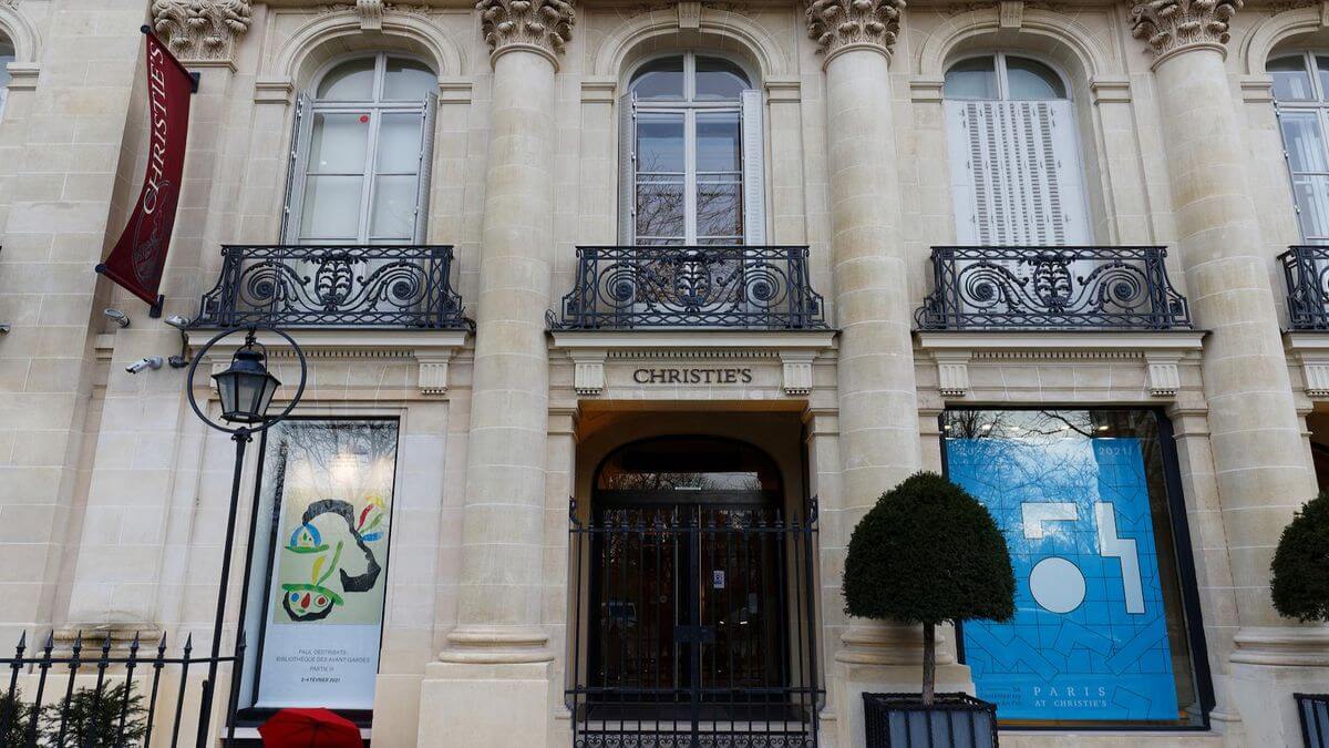 Exterior of Christie's auction house with stone columns, ornate black balcony railings, and poster displays in windows on either side of the entrance.