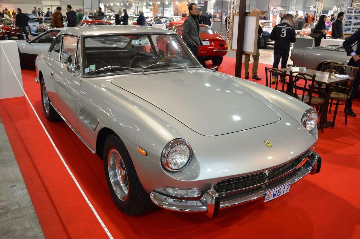 Silver classic Ferrari coupe displayed on red carpet at a car show with people and other cars in the background.
