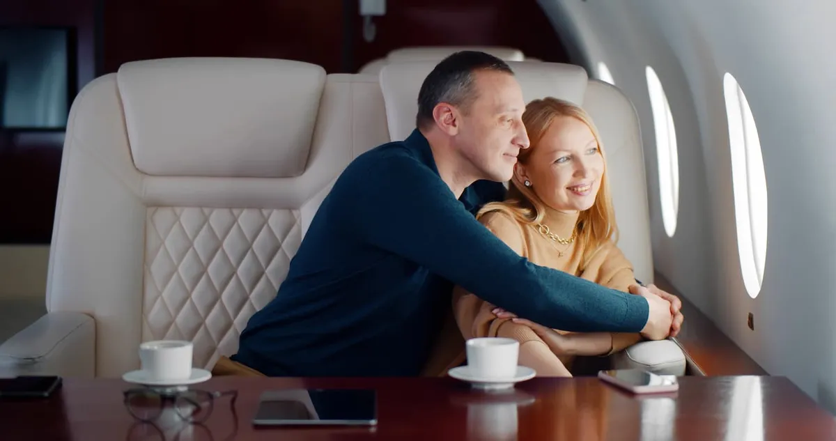 Couple sitting closely together and smiling, looking out the window of a private jet with beige leather seats and a wooden table holding cups and tablets.