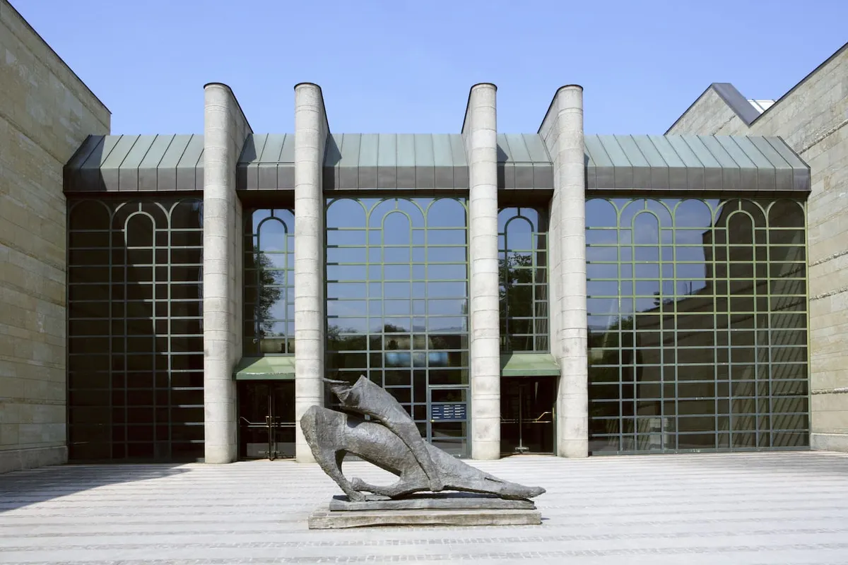 Modern building facade with large glass windows, four cylindrical stone columns, and an abstract stone sculpture in front on a striped plaza.