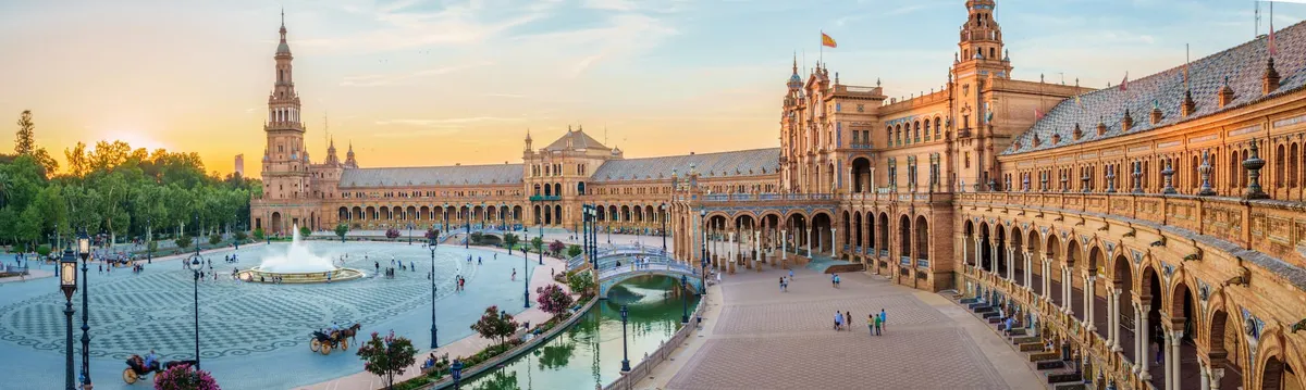 Wide view of Plaza de España in Seville, Spain, with ornate buildings, a central fountain, a canal with bridges, and people walking.