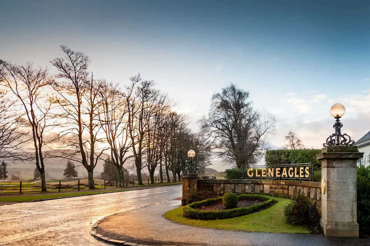 Curved road at entrance to Gleneagles with bare trees lining one side and a stone sign with lamps.