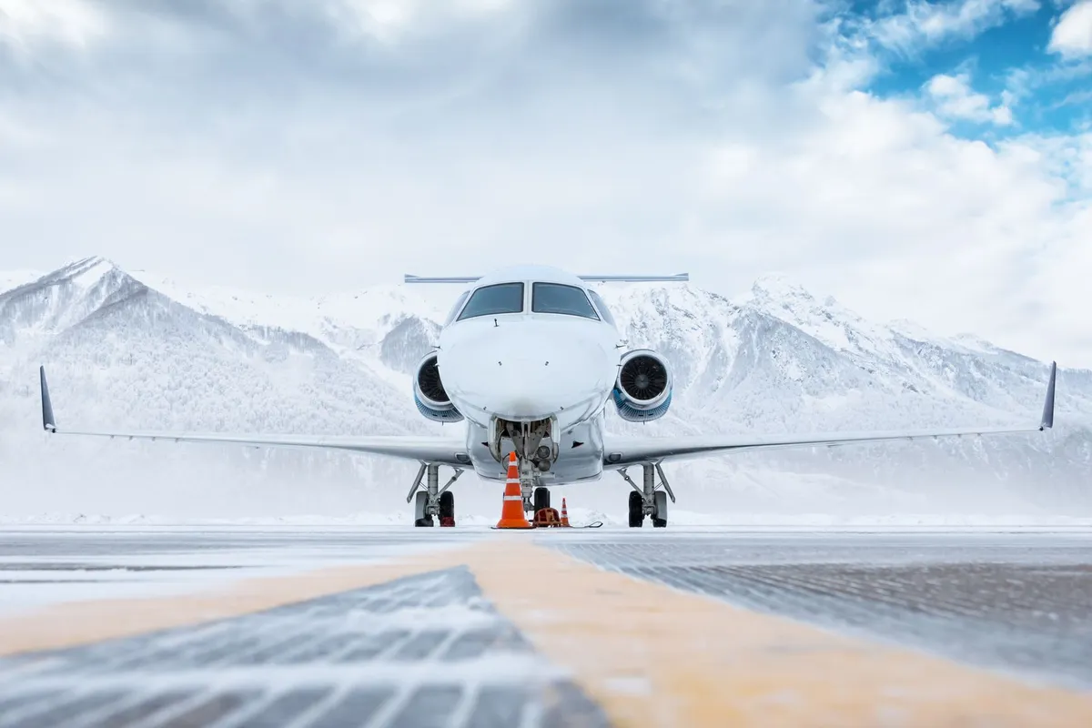 Front view of a white airplane parked on a snowy runway with snow-covered mountains and a cloudy blue sky in the background.