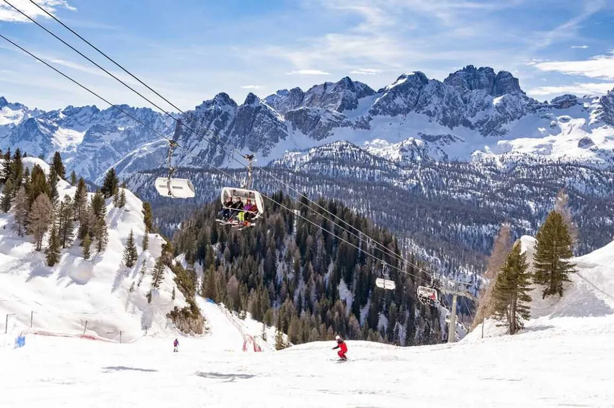 Skiers on snowy slope with chairlift and pine trees against a backdrop of snow-covered mountain peaks under a partly cloudy sky.