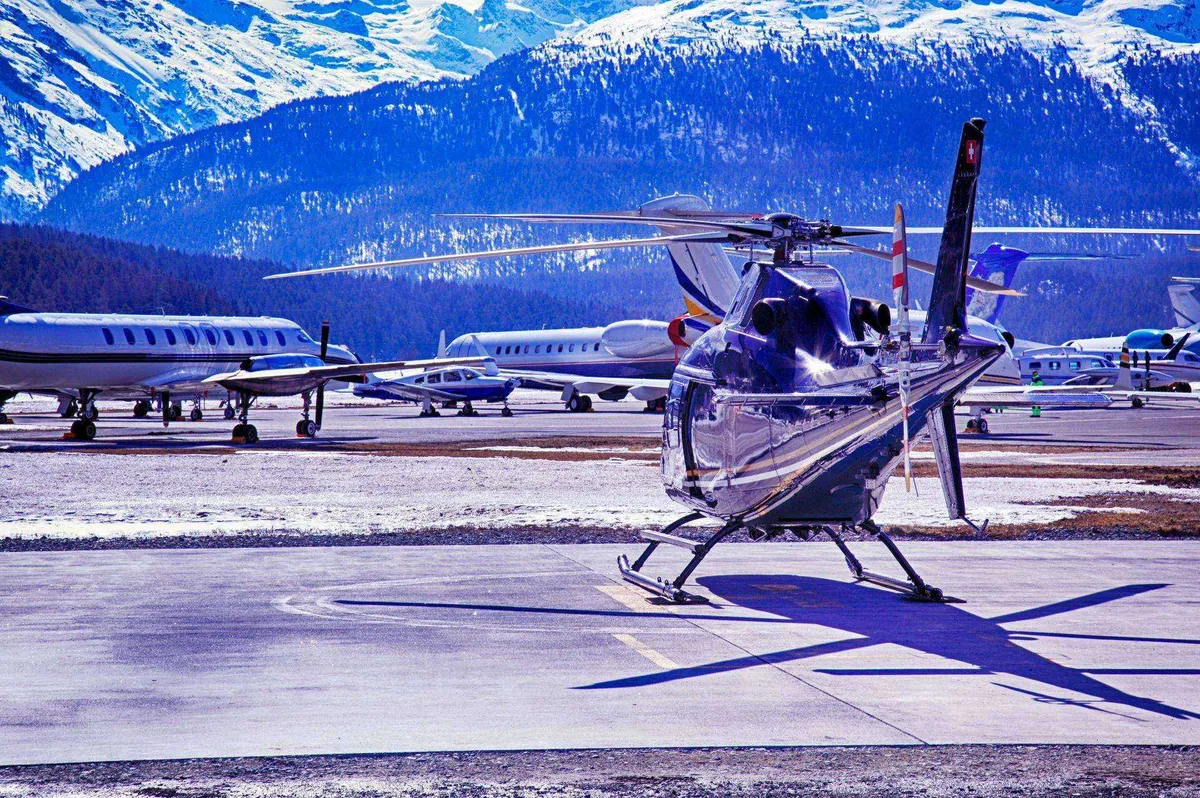 Helicopter parked on a snowy airfield with several small airplanes and snow-covered mountains in the background.