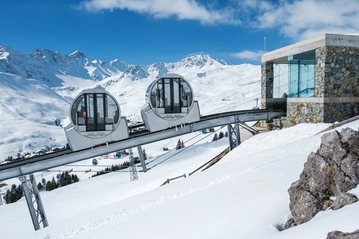 Two modern cable cars on a track ascending a snowy mountain landscape with a stone station building and snow-covered peaks in the background.