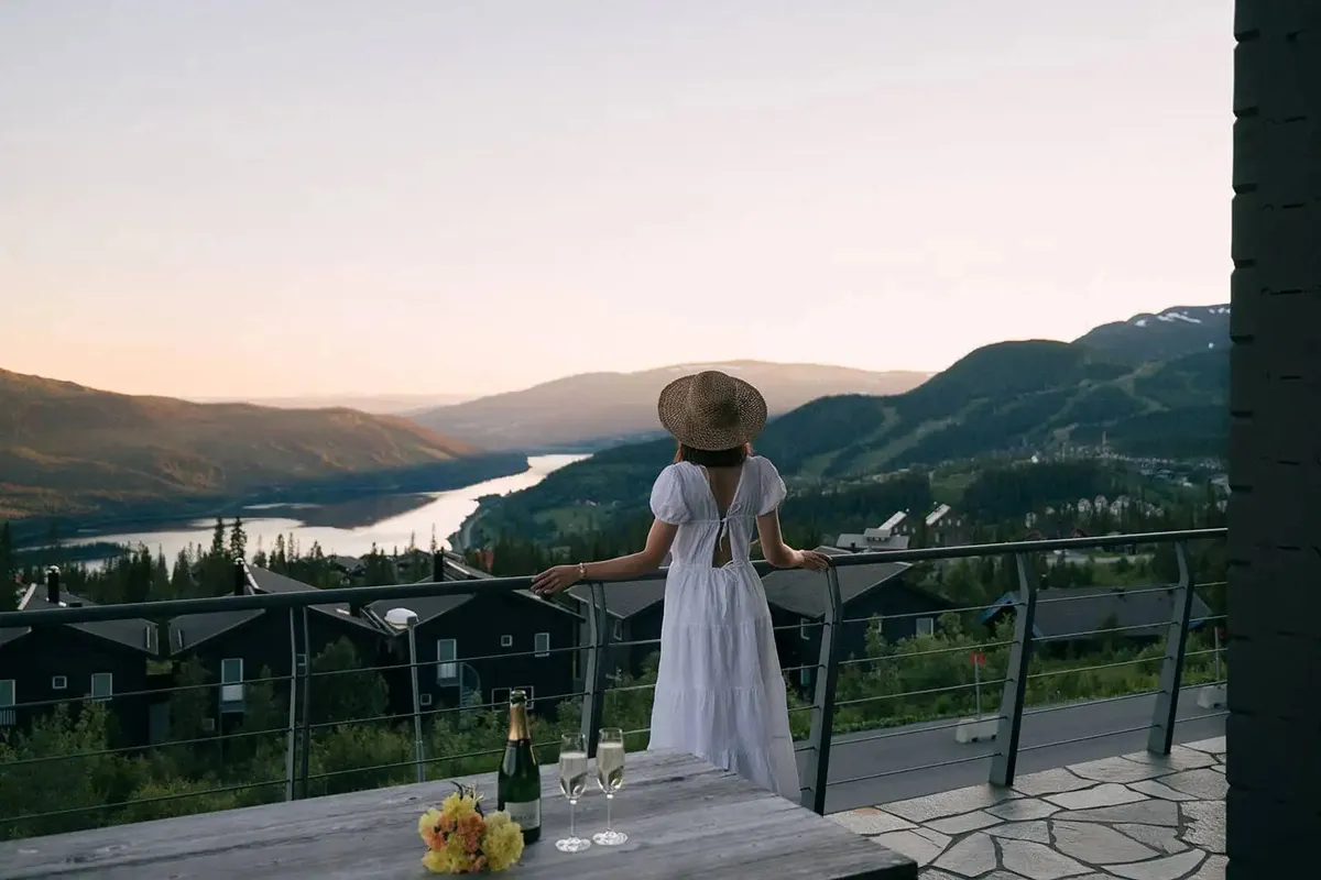 Woman in white dress and straw hat standing on balcony overlooking river valley and mountains at sunset.