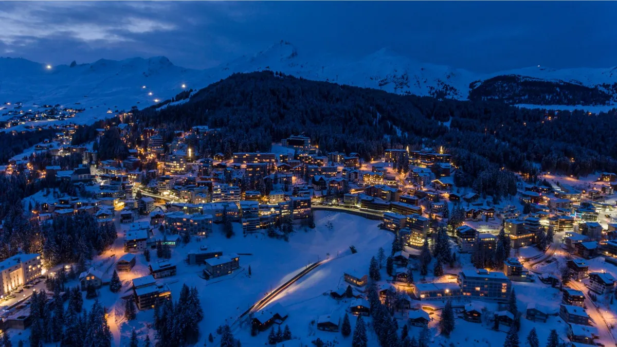Arosa, Switzerland town illuminated at twilight with snow-covered buildings and surrounding forested mountains.