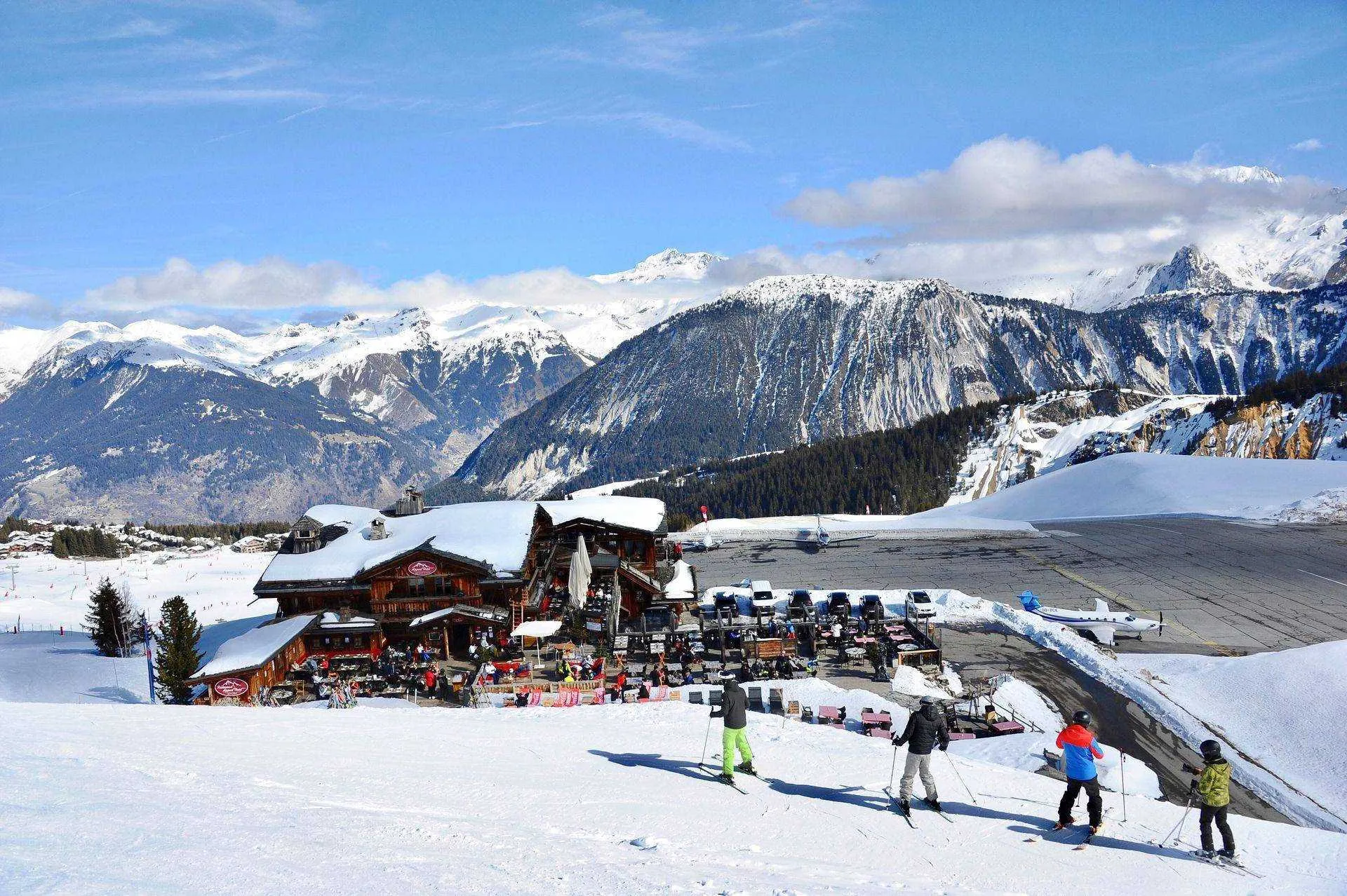 Four skiers on a snowy slope near a wooden mountain lodge with outdoor seating, surrounded by snow-covered mountains under a clear blue sky.