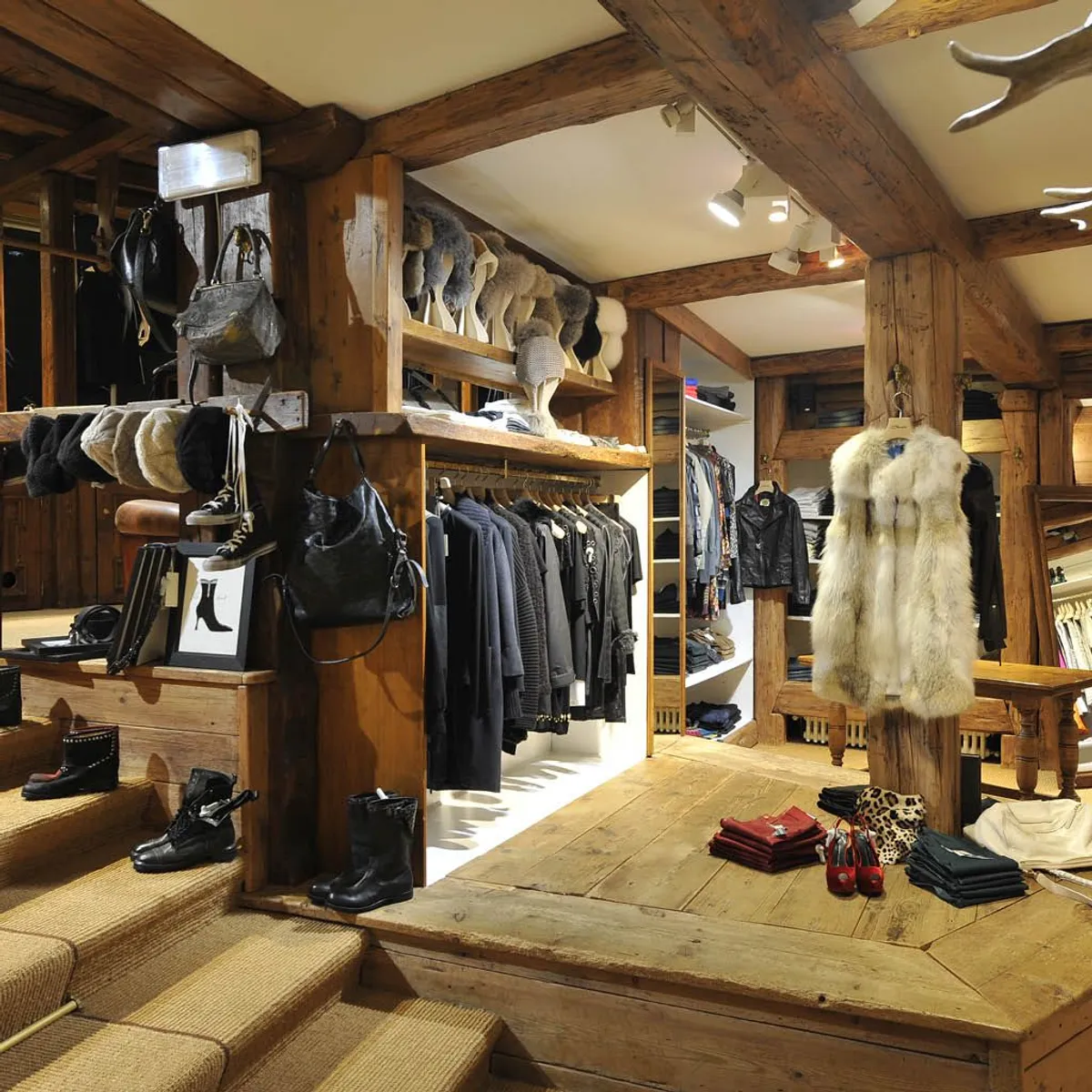 Cozy clothing store interior with wooden beams, hangers of black coats, fur hats on mannequin heads, boots on carpeted stairs, and folded clothes with a fur vest on display.