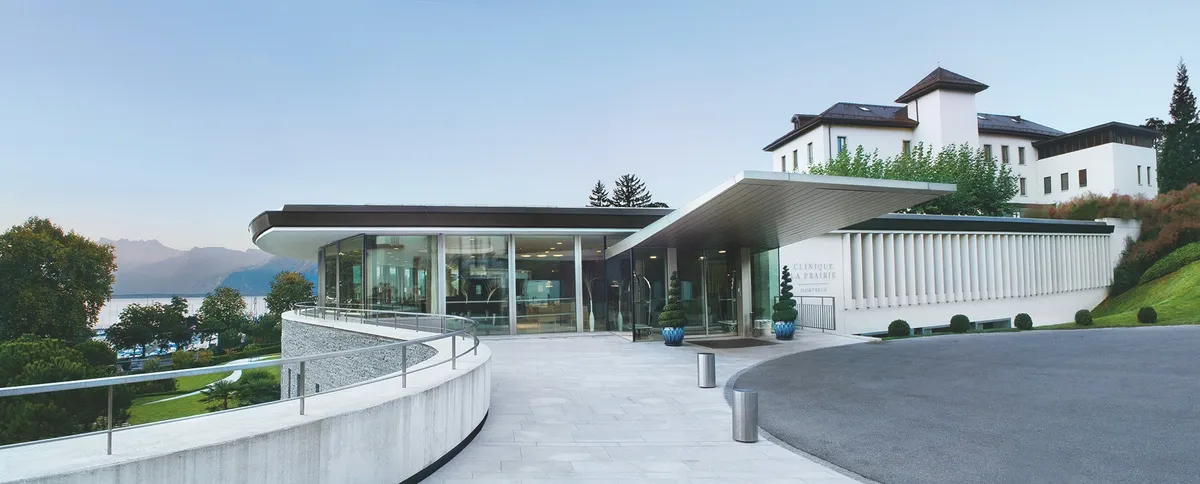 Modern wellness center with glass walls and a curved walkway, surrounded by greenery and mountains in the background.
