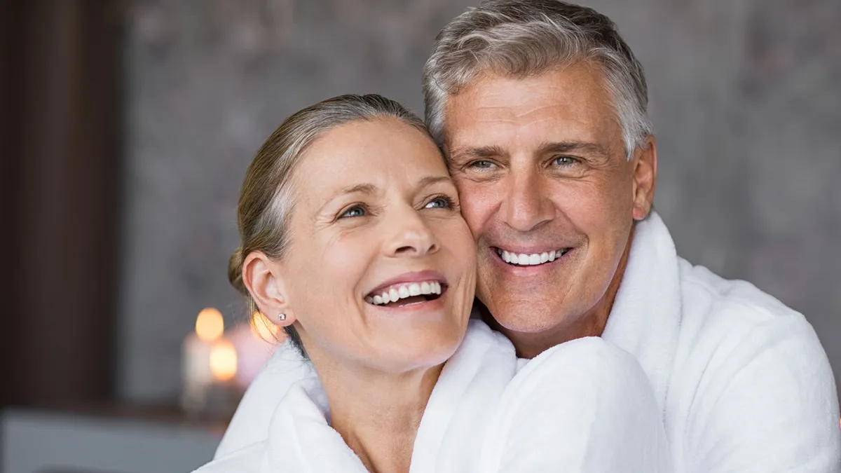 Smiling middle-aged couple in white robes embracing indoors with blurred candles in the background.
