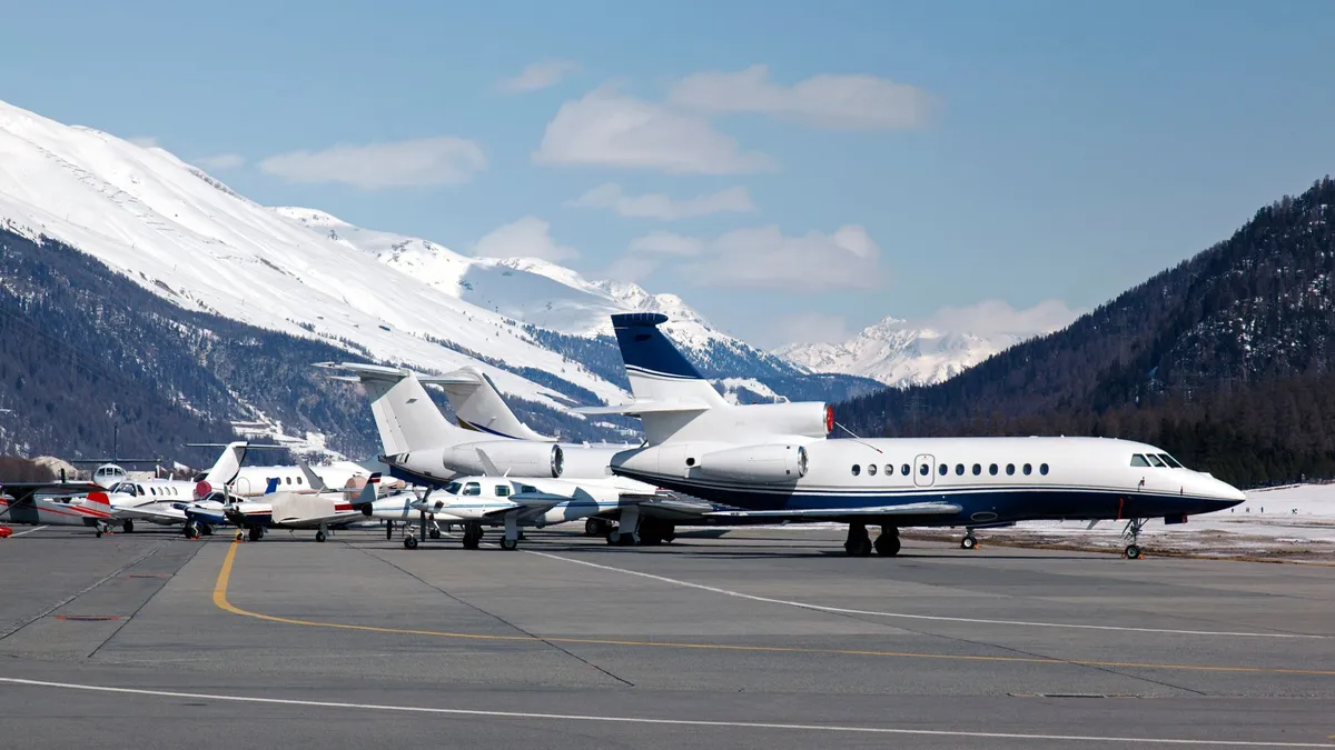 Several private jets and small planes parked on a snowy mountain airport tarmac with snow-covered peaks in the background.