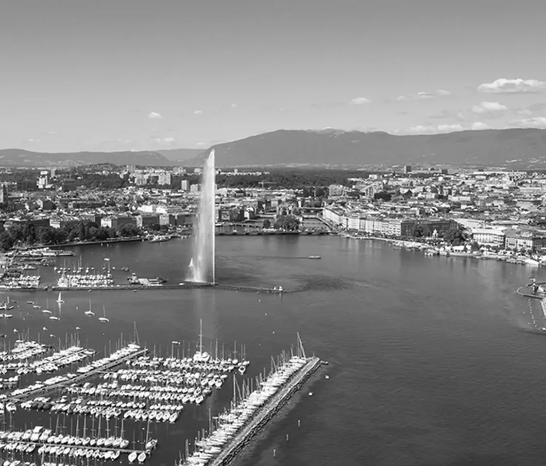 Вид с воздуха на пристань с лодками и большим фонтаном, брызгающим посреди озера, окруженного городскими зданиями и горами.