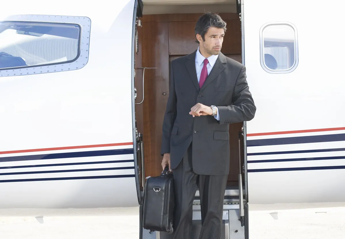Businessman in dark suit stepping off a private jet, holding a briefcase and checking his wristwatch.