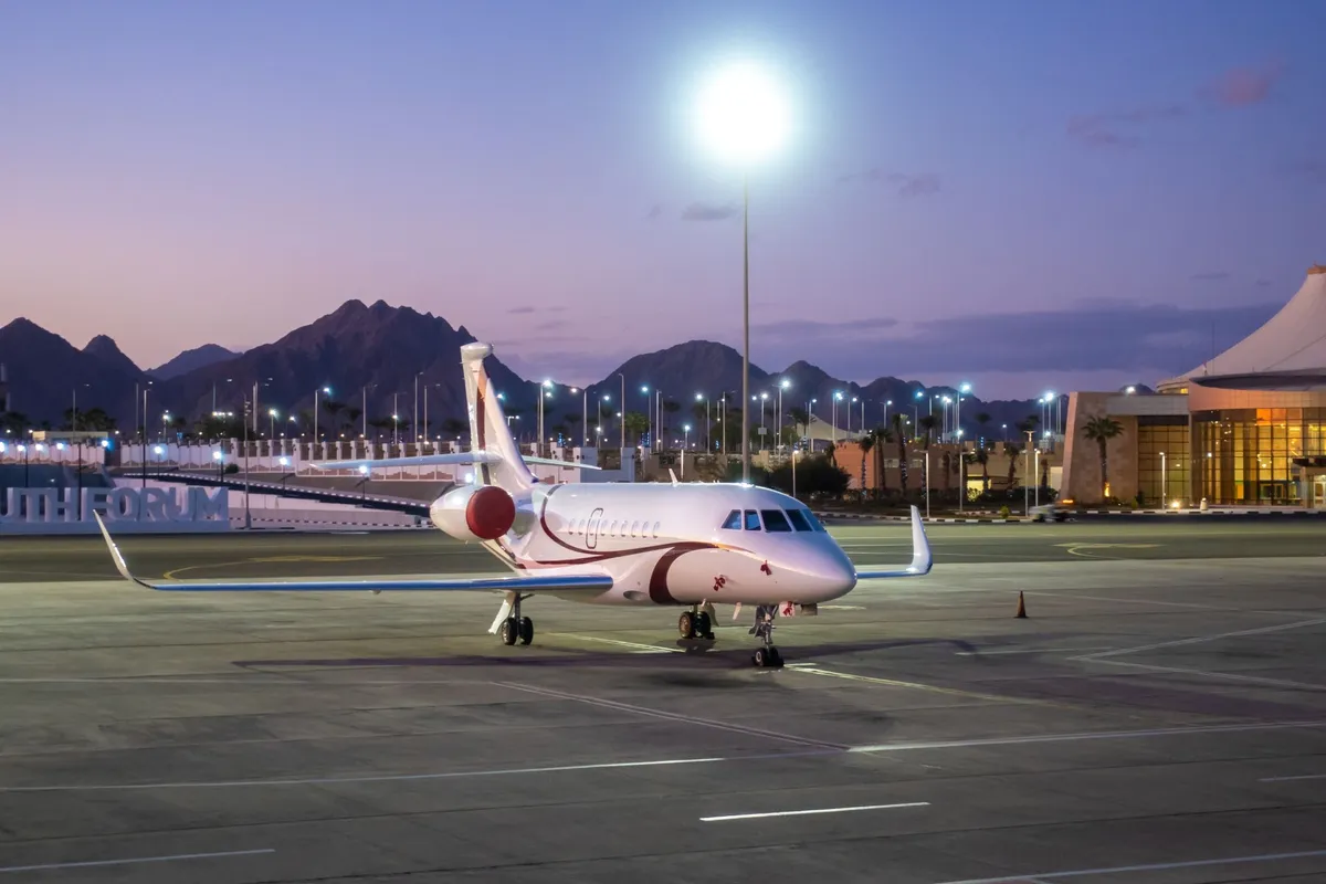 Corporate jet parked on airport tarmac at dusk with distant mountains and airport buildings in the background.