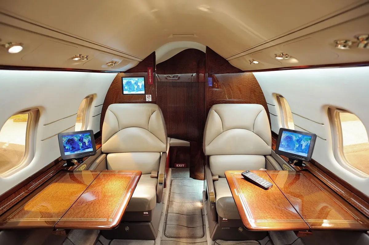 Luxurious private jet cabin with two cream leather seats facing wood-finish tables and small monitors by the windows.