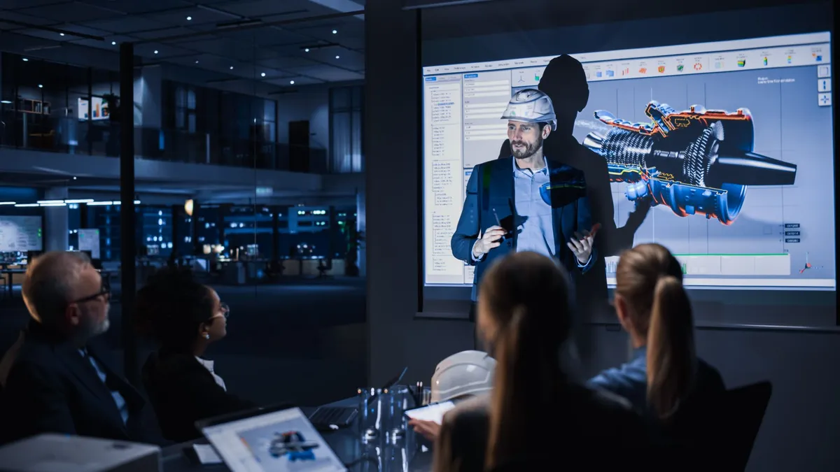 Man in hard hat giving a technical presentation of a 3D engine model to a seated team in a dim office.