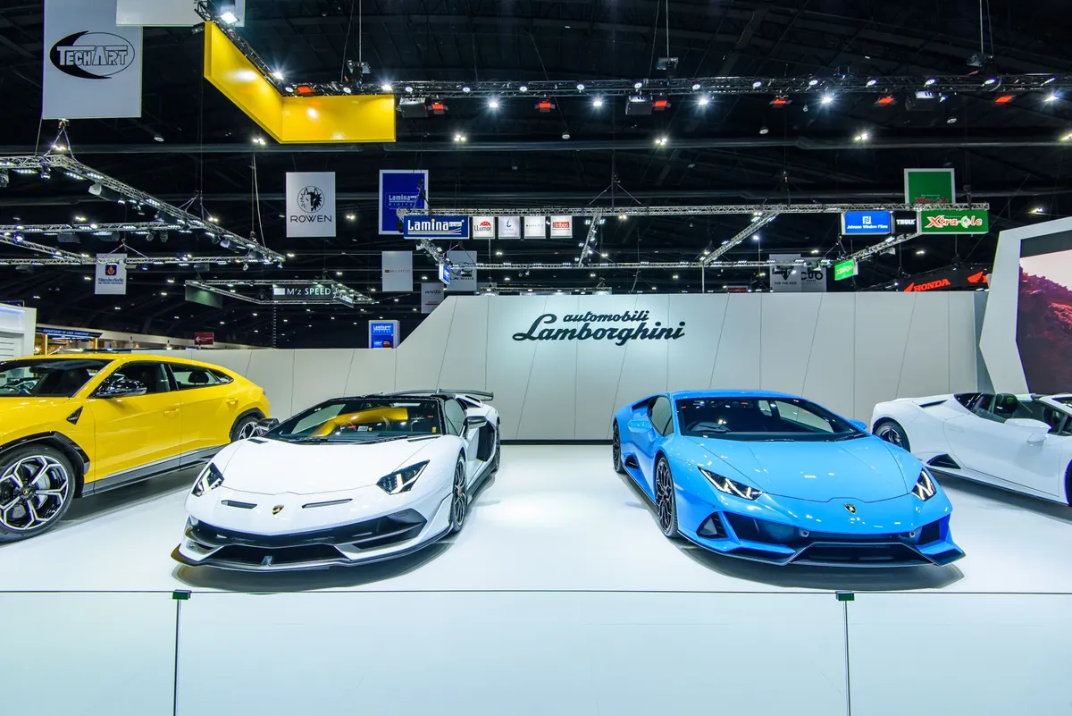 White and blue Lamborghini sports cars displayed at an automobile show with Lamborghini logo on the back wall.