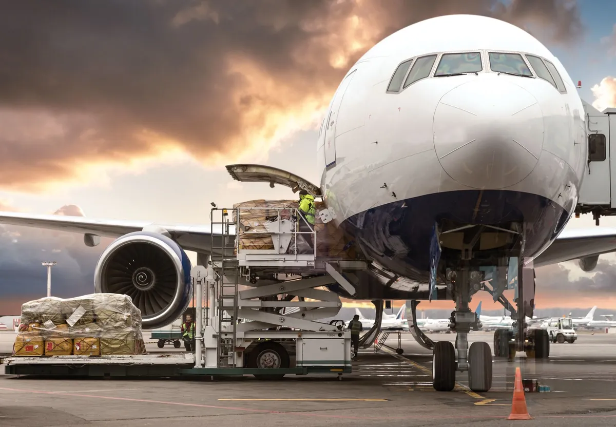 Cargo plane being loaded with pallets of goods by workers using a scissor lift at an airport during sunset.