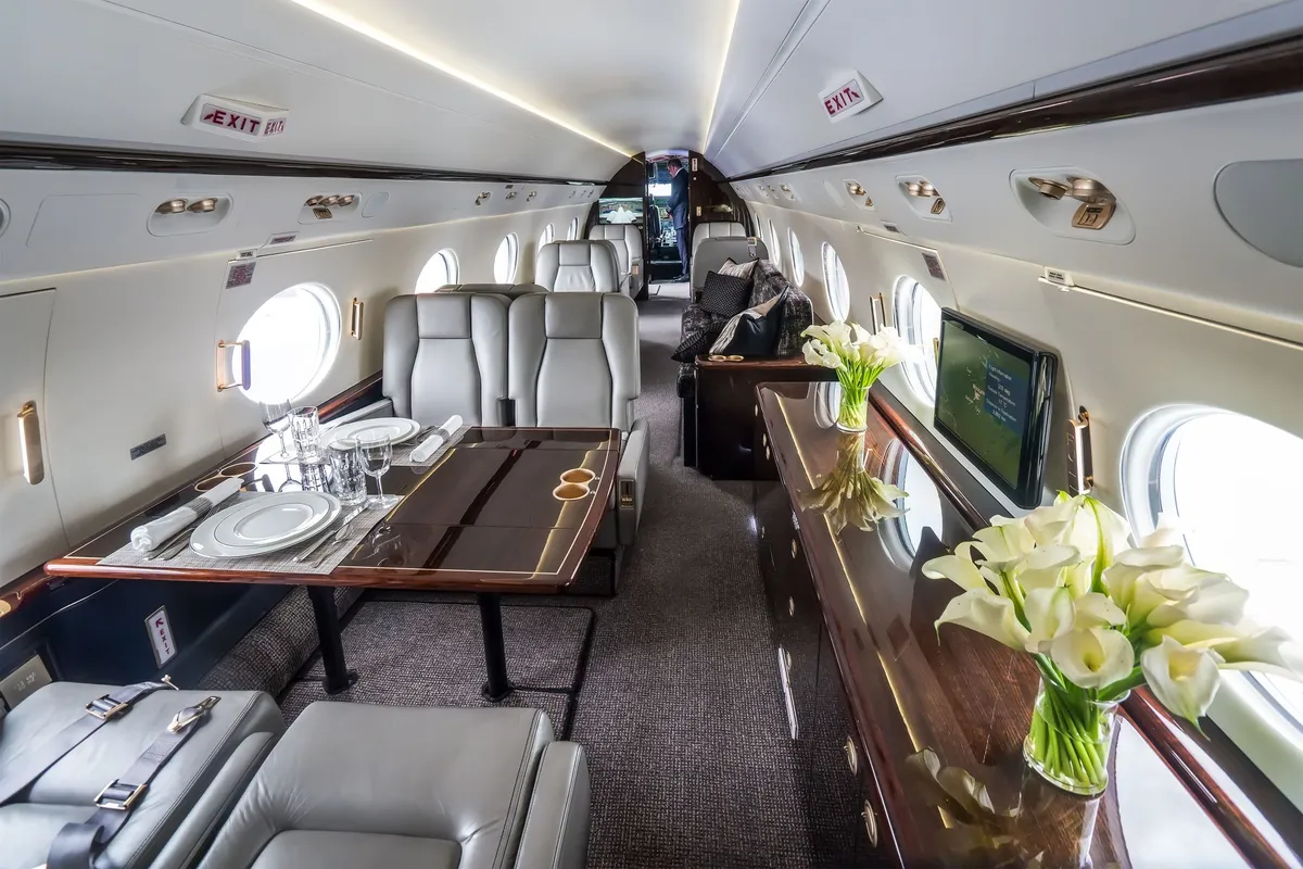 Luxurious private jet interior with leather seats, a dining table set with plates and glasses, a wooden cabinet with a flower vase, and a man standing near the cockpit.