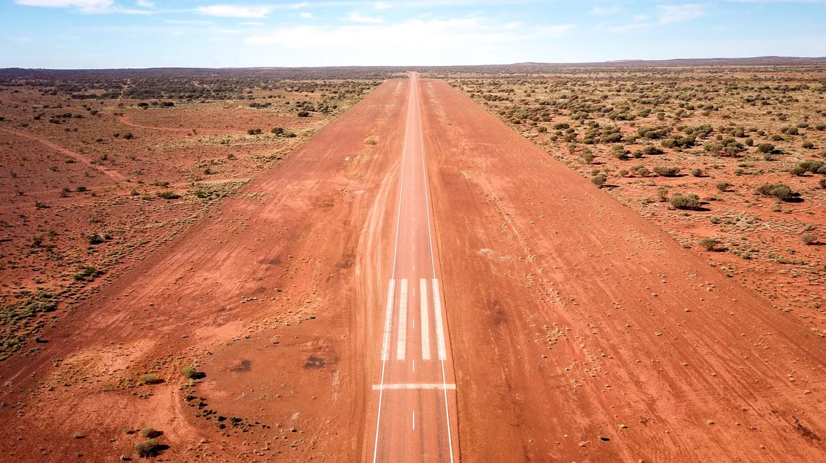 Aerial view of a narrow red dirt runway extending into the distance through a dry, sparsely vegetated landscape.