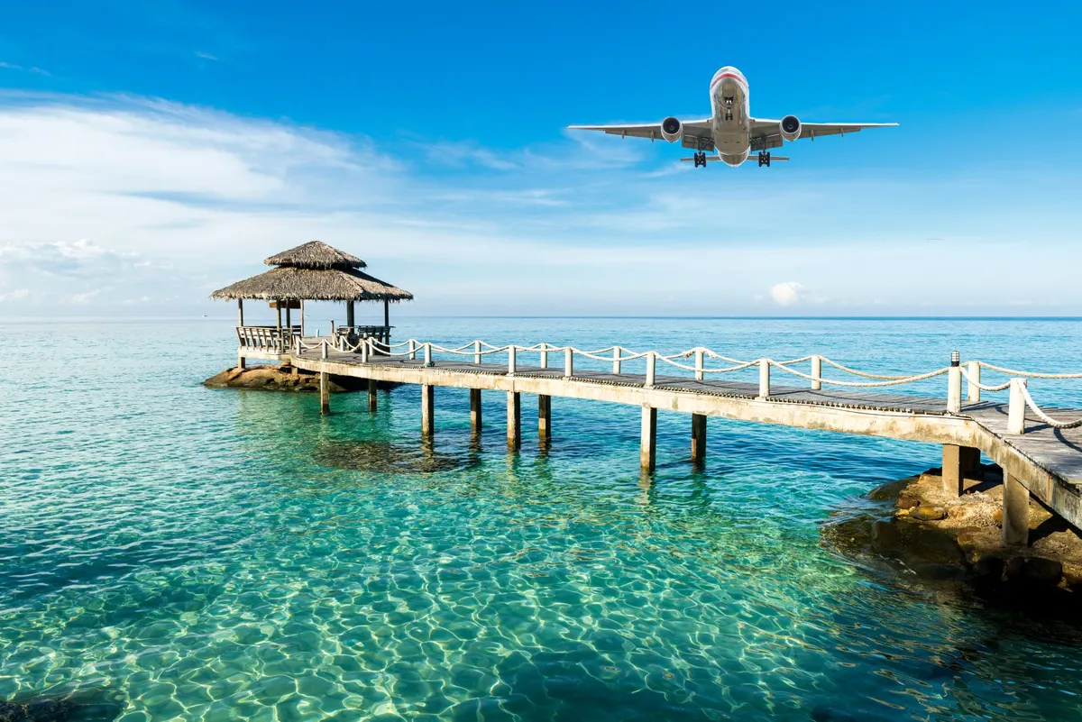 Airplane flying low over a wooden pier with a thatched-roof shelter on clear turquoise water under a blue sky.