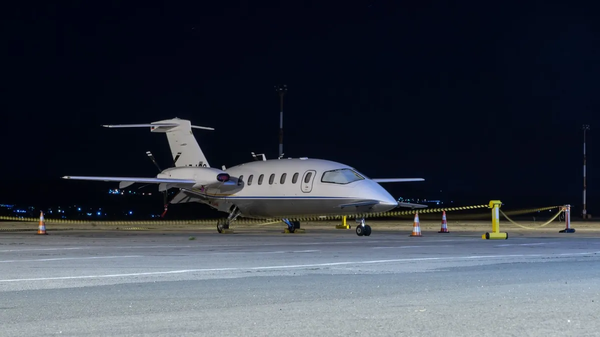 Side view of a small white private jet parked on a tarmac at night with safety cones and barriers around it.