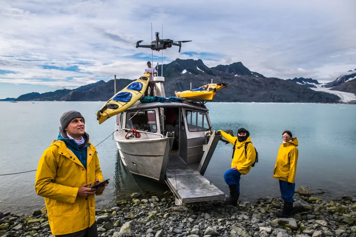 Three people in yellow jackets standing by a small boat with kayaks on top near a rocky shore and mountains under a cloudy sky.
