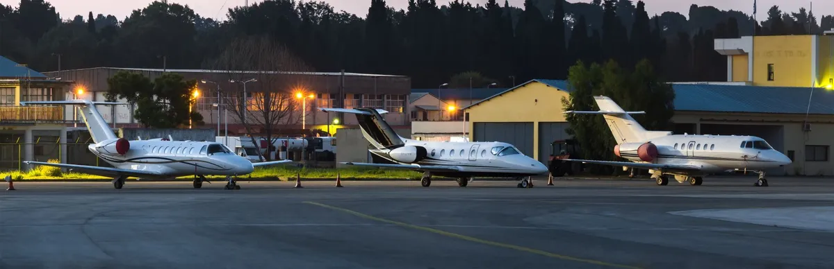 Three private jets parked in a row on an airport tarmac at dusk with buildings and trees in the background.