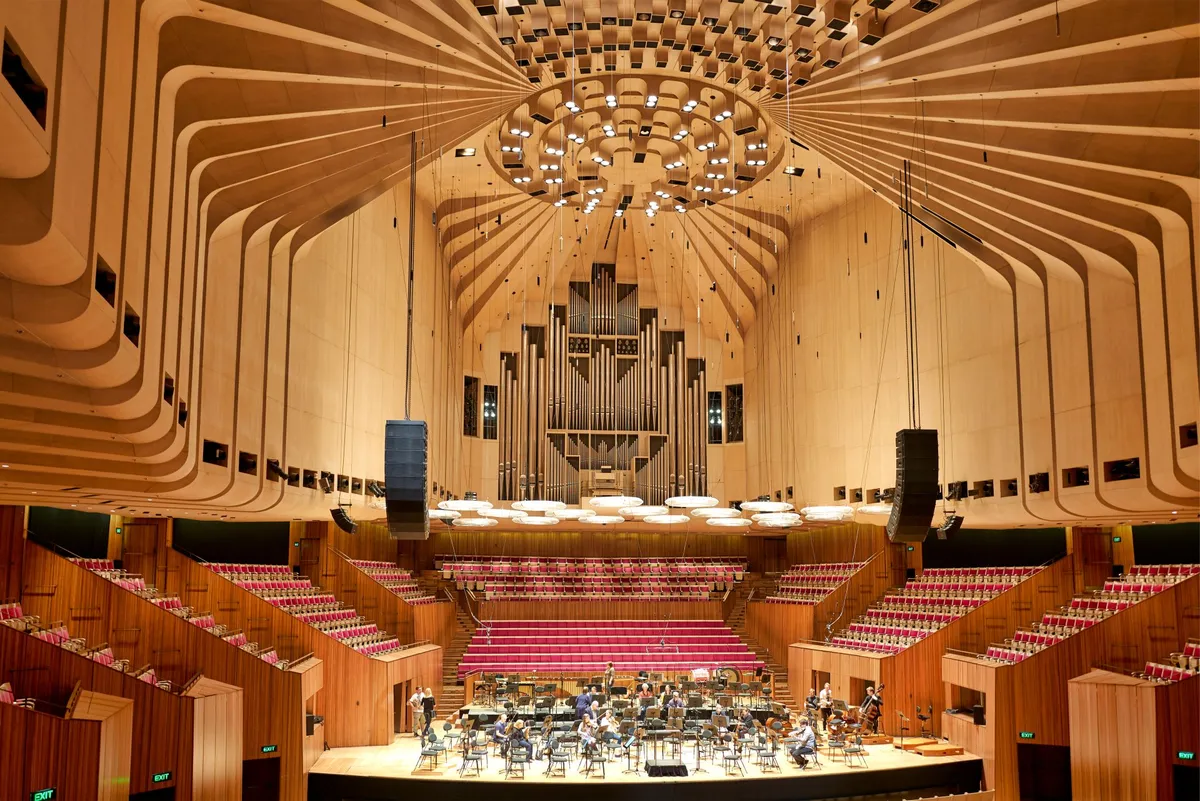 Wide view of a concert hall with wooden walls, red seats, a ceiling organ, and musicians setting up on stage.