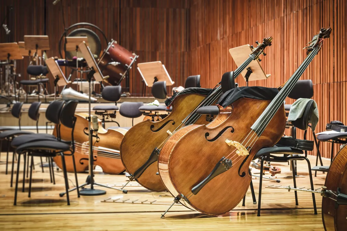 Cellos resting on chairs in an empty orchestra rehearsal room with music stands and percussion instruments in the background.