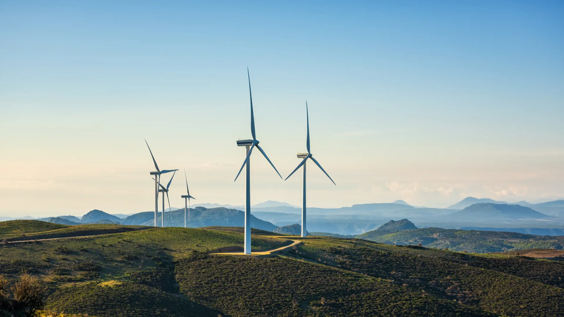 Several wind turbines on green hills with distant blue mountains under a clear sky.