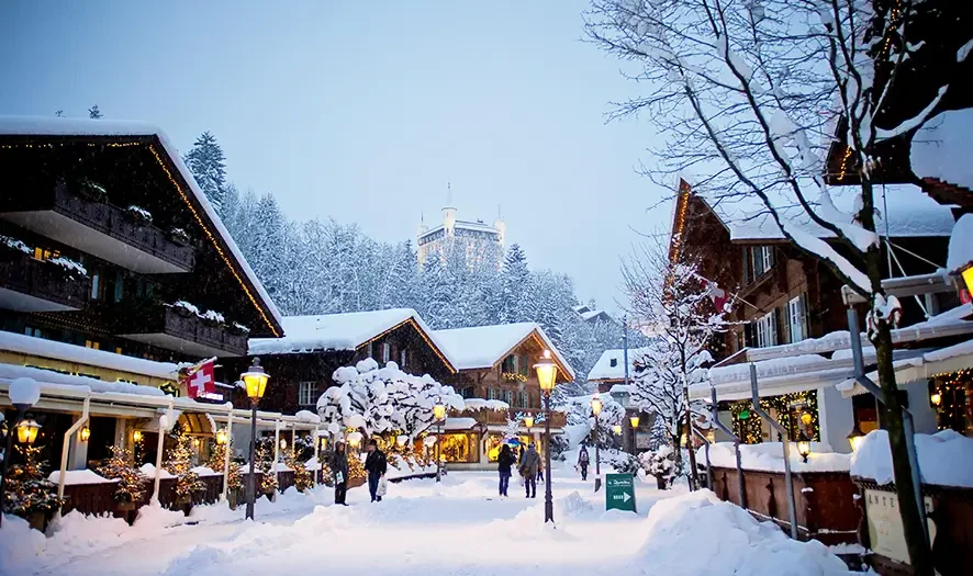 Snow-covered alpine village street with wooden chalets decorated with lights and people walking under a pale blue sky.