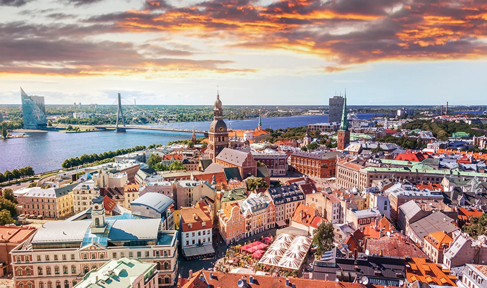 Aerial view of Riga cityscape with colorful buildings, churches, river, and a dramatic sunset sky.