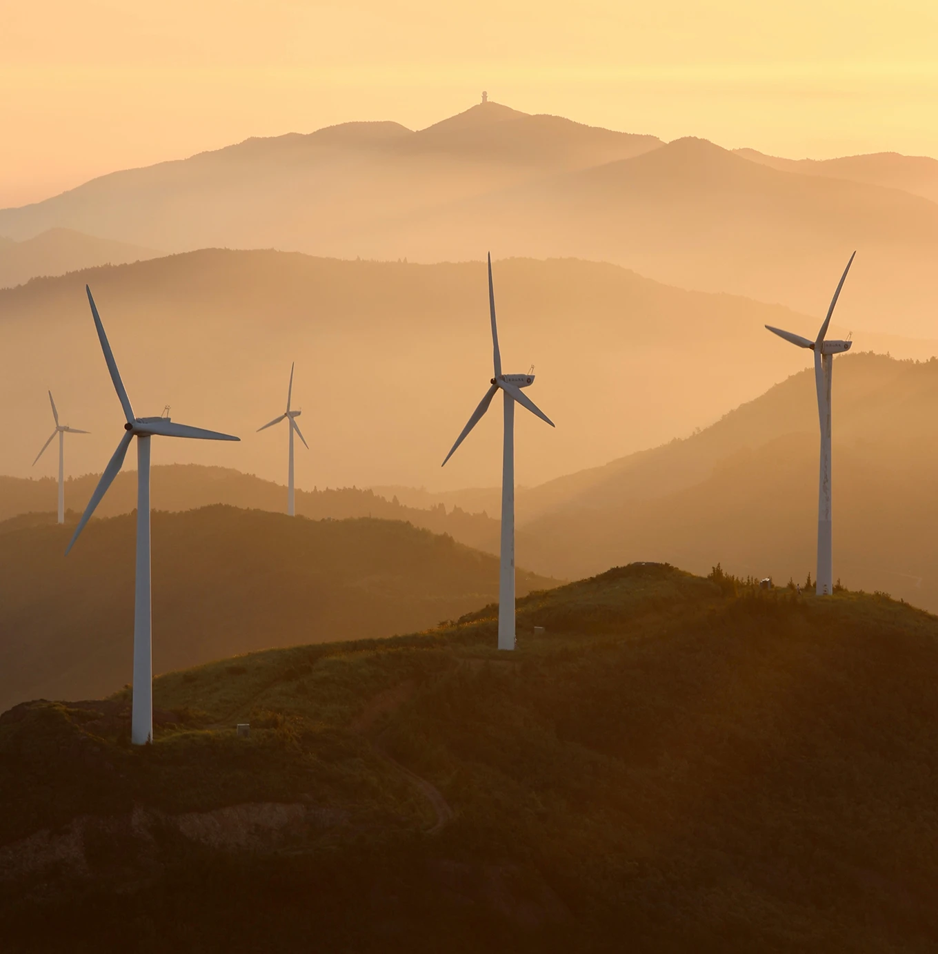 Wind turbines standing on green rolling hills with layered mountain silhouettes in warm golden sunset light.