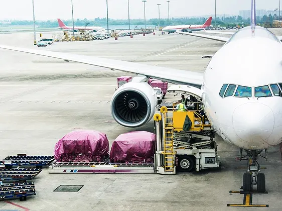 Ground crew loading luggage covered in purple plastic onto a white passenger airplane at an airport tarmac.