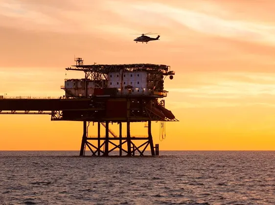 Helicopter flying above an offshore oil rig at sunset with calm ocean water.