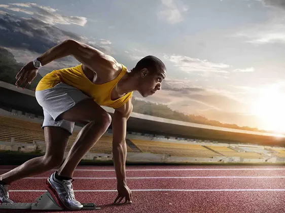 Athletic runner in a yellow tank top and gray shorts poised at the starting blocks on a track at sunset.